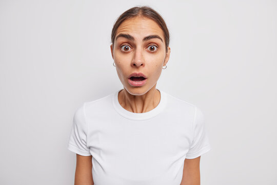 Portrait of shocked young European woman with combed dark hair gasps from wonder being scared of something dressed in casual t shirt isolated over white background. People and emotions concept