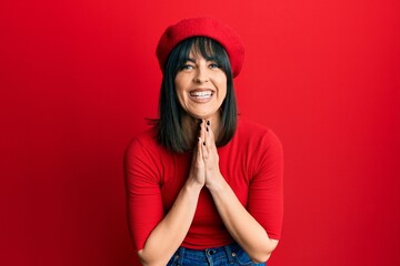 Young hispanic woman wearing french look with beret praying with hands together asking for forgiveness smiling confident.