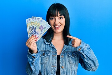 Young hispanic woman holding 100 romanian leu banknotes smiling happy pointing with hand and finger - obrazy, fototapety, plakaty