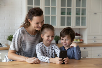 Happy caring millennial mother controlling small children son daughter using cellphone applications, playing online mobile video games, watching funny photo video content in social network in kitchen.