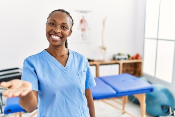 Black woman with braids working at pain recovery clinic smiling cheerful offering palm hand giving assistance and acceptance.