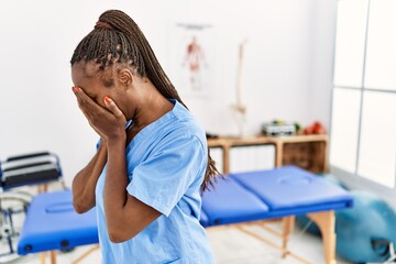 Black woman with braids working at pain recovery clinic with sad expression covering face with hands while crying. depression concept.