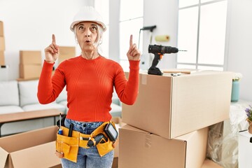 Middle age grey-haired woman wearing hardhat standing at new home amazed and surprised looking up and pointing with fingers and raised arms.