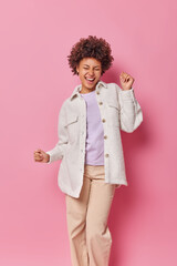 Carefree overjoyed woman dances and laughs happily has upbeat mood dressed in fashionable clothes isolated over pink background. Stylish curly haired female model moves with rhythm of music.