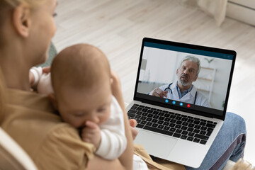 Caring young mother holding sleeping baby, using laptop, making video call to mature man doctor at home, senior pediatrician consulting loving mom online, telemedicine and child healthcare concept
