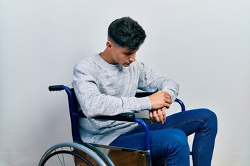 Young hispanic man sitting on wheelchair checking the time on wrist watch, relaxed and confident