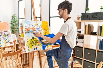 Young hispanic artist man smiling happy drawing using paint roller at art studio.