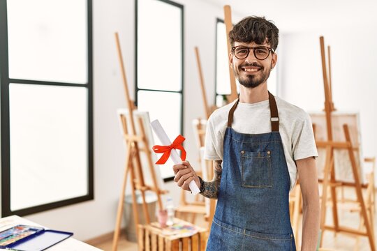 Young hispanic artist man smiling happy holding diploma at art studio.