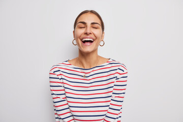 Indoor shot of happy woman keeps eyes clsed smiles broadly shows teeth laughs at something positive dressed in casual striped jumper isolated over white backgroud. People and emotions concept