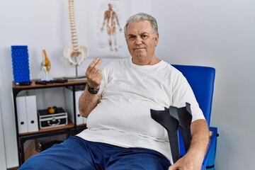 Senior caucasian man at physiotherapy clinic holding crutches doing money gesture with hands,...