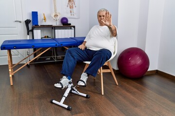 Fototapeta premium Senior caucasian man at physiotherapy clinic using pedal exerciser doing stop sing with palm of the hand. warning expression with negative and serious gesture on the face.