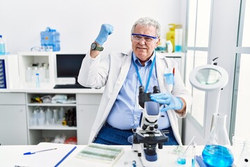 Senior caucasian man working at scientist laboratory angry and mad raising fist frustrated and furious while shouting with anger. rage and aggressive concept.
