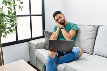 Young handsome man with beard using computer laptop sitting on the sofa at home sleeping tired dreaming and posing with hands together while smiling with closed eyes.