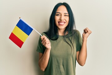 Young hispanic girl holding romania flag screaming proud, celebrating victory and success very...