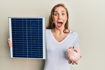 Young blonde woman holding photovoltaic solar panel and piggy bank celebrating crazy and amazed for...