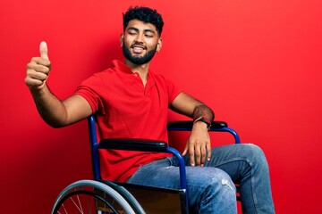 Arab man with beard sitting on wheelchair looking proud, smiling doing thumbs up gesture to the side