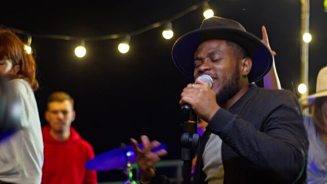 Beautiful young african american man singing song on a rooftop party. Friends enjoying rooftop party music, celebrating weekend reunion gathering.