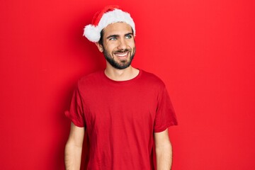 Young hispanic man wearing christmas hat looking away to side with smile on face, natural...