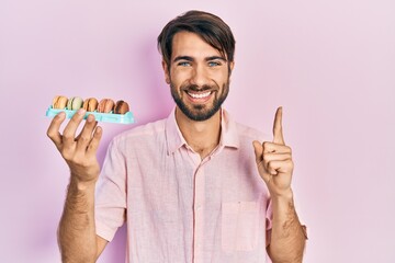 Young hispanic man holding delicious macrons pastries smiling with an idea or question pointing finger with happy face, number one