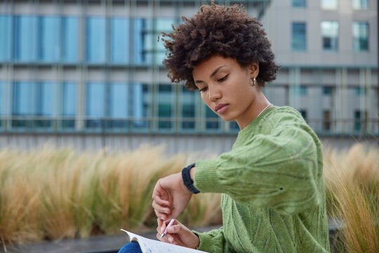 Serious curly woman writes notes in notepad learns material prepares for exams checks time on wristwatch wears casual green jumper poses outdoors against city building involved in learning process