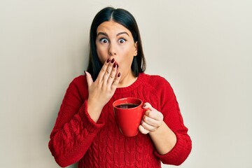 Beautiful brunette woman drinking a cup of black coffee covering mouth with hand, shocked and...