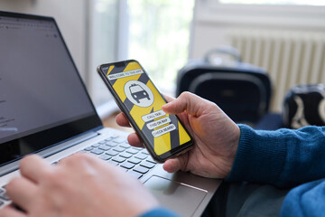 Closeup of a man working on laptop and using mobile phone to call a taxi while waiting to travel with suitcase in background.