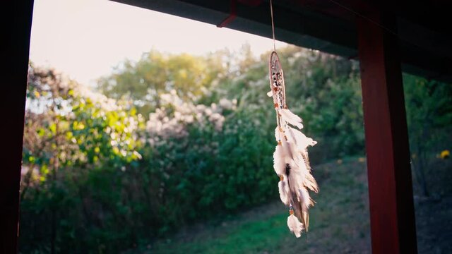 A beautiful cinematic shot of a knitted dreamcatcher with feathers and stones hangs on the veranda against the backdrop of the sun and a garden.