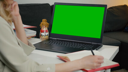 Green Screen with Color Laptop Display. Businesswoman sitting at table takes notes in notebook from laptop,