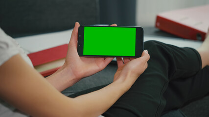 Young Woman sitting on sofa while looking at movies using phone in horizontal mode with mock up green screen chroma key display.
