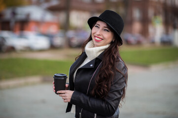 A joyful white female model in a black leather jacket, wearing a black hat, looks away with a smile on her face. Street photo of a beautiful Caucasian woman drinking coffee and laughing.