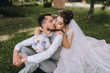 A beautiful, sweet bride in a dress hugs her back, kisses a stylish, bearded, smiling groom in a gray suit and white shirt, sitting on the green grass. Wedding portrait, photo of newlyweds.