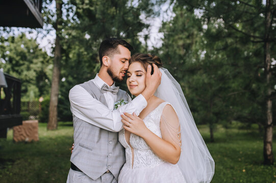 A young, stylish groom in a gray checkered suit and a beautiful, sweet bride in a white dress are hugging in the woods in nature. Wedding portrait, photo of newlyweds.