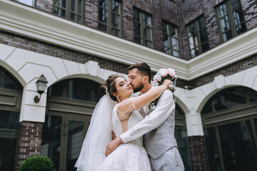 Naklejka premium A beautiful bride in a white dress with a long veil and a stylish bearded groom in a gray suit with a bow tie are hugging against the backdrop of architecture, red brick houses with windows.