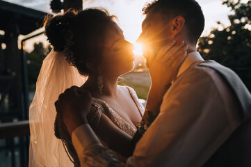 Stylish groom in a gray vest and shirt with a bow tie kisses a beautiful, sweet bride in a white dress against the background of the sun. Wedding photo of the newlyweds at sunset close-up.