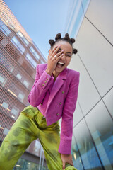 View from below of overjoyed female model keeps hand on face feels very happy wears stylish pink jacket and green trouses spends free time in urban place expresses positive emotions. Youth style - obrazy, fototapety, plakaty