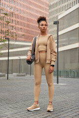 Full length shot of thoughtful slim woman in sportswear carries yoga mat going to practice has motivated expression poses in urban place. Sporty female has regular training every day. Wellness