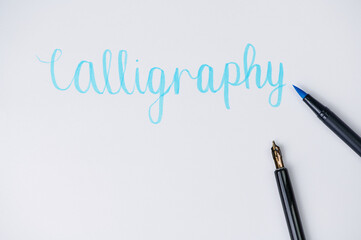 the word Calligrahpy written on a white paper beside a brush pen and fountain pen, focus on the blue felt pen