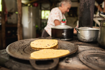 Primer plano de tortillas de maíz en un comal de una cocina de metal a la leña con una anciana de...