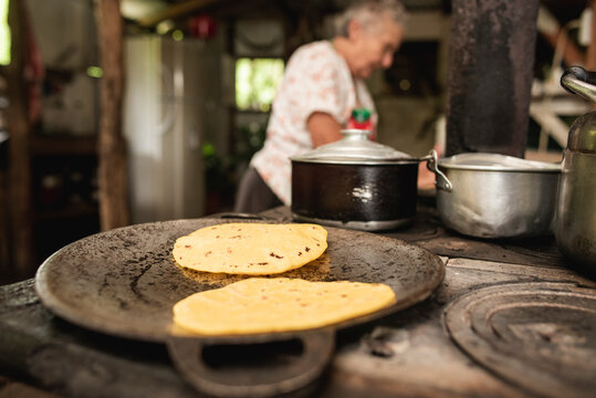 Primer plano de tortillas de ma&iacute;z en un comal de una cocina de metal a la le&ntilde;a con una anciana de fondo