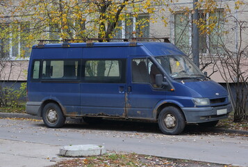 An old blue rusty minibus is parked in the courtyard of a residential building, Dybenko street, St....
