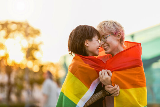 Young non binary couple in love hugging under a rainbow flag 
