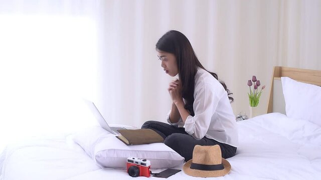 Young Asian woman sitting on the bed and preparing for trip, using a laptop computer to searching information for travel is opening a country after quarantine in hotel during the coronavirus outbreak.