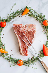 raw t-bone steak in the center of a Christmas wreath of rosemary thyme cherry tomatoes and bell peppers on a white marble surface. christmas dinner concept
