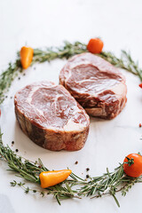 raw two ribeye steaks in the center of a Christmas wreath of rosemary thyme cherry tomatoes and bell peppers on a white marble surface. christmas dinner concept for two