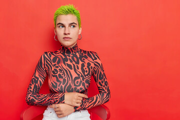 Horizontal shot of thoughtful hipster girl has bright dyed green short hair looks with pensive expression dressed in casual clothes poses on chair against red background copy space on right.