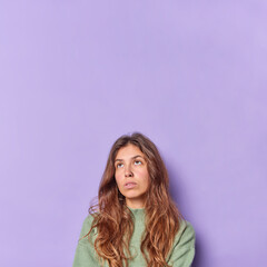 Naklejka premium Vertical image of thoughtful long haired woman has bored unhappy expression focused above thinks about something wears casual jumper isolated over purple background with blank space for your text