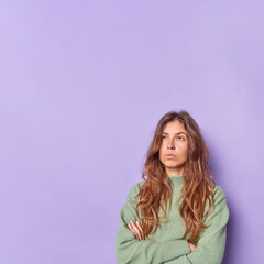 Thoughtful beautiful young woman keeps arms folded concentrated above thinks about something wears casual jumper isolated over purple background with copy space for your advertising content.
