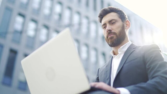 young bearded business man freelancer sitting on bench and working with laptop in city park on modern urban street background outdoors downtown Male enjoy outside Focused entrepreneur browsing typing