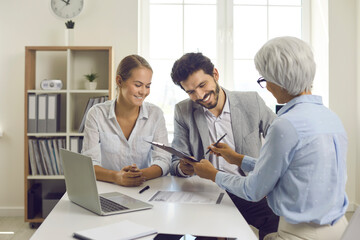 Fototapeta premium Happy young married couple signs documents after successful consultation with female banking agent. Concept of insurance contract, budget planning, loan agreement or contract of sale of real estate.
