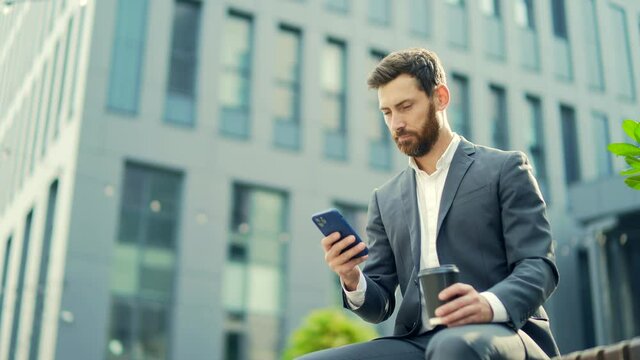 Handsome bearded businessman sitting on bench with cup coffee use mobile phone browse smartphone in hands reads the news of using a smart. Downtown. Outside a modern urban street background. City park